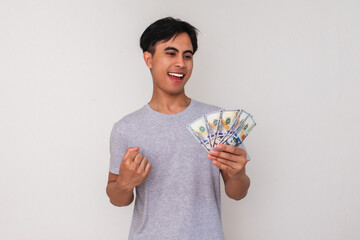 Portrait of a happy young Asian man holding cash and clenching his fist in celebration, smiling...