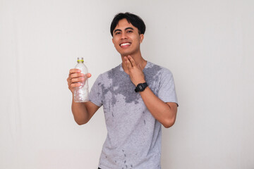 Young Asian man smiling and holding a water bottle with a satisfied expression, sweating after...