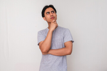 Portrait of a young Asian man thinking seriously with hand on chin and eyes looking up, isolated on...