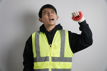 Energetic young Asian man in a safety vest and gloves, shouting and raising his fist in a gesture of triumph, protest, or excitement.