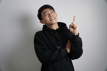 A contemplative young Asian man in a black hoodie looks up and points his index finger, suggesting an idea or a thought.