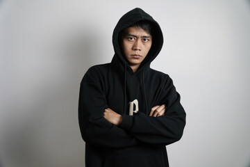A serious young Asian man wearing a black hooded sweatshirt stands with his arms crossed, looking directly at the camera with a stern expression.