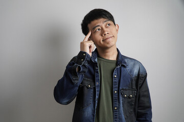 A thoughtful young Asian man in a denim shirt points his finger to his temple