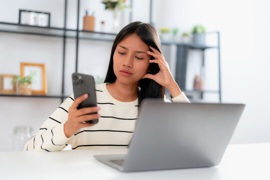 Young asian woman sitting at desk with laptop, looking stressed and confused while checking her smartphone, concept of online problem, bad news, finance issue, or work stress at home
