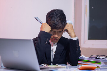 Asian businessman sitting at desk, looking thoughtful and stressed. There are many financial documents being analyzed about banking, investing, borrowing, selling, buying.