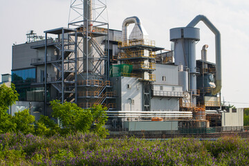A towering industrial chimney rises above a complex of factory buildings under a cloudy sky