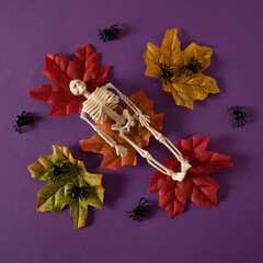A human skeleton rests on fallen autumn leaves, surrounded by small black spiders.