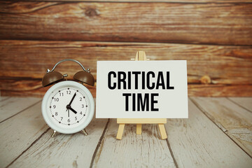 Critical Time text with easel and alarm clock on wooden background