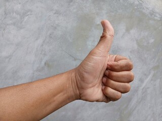 A man's hand shows a thumbs up gesture meaning OK with a gray wall in the background.