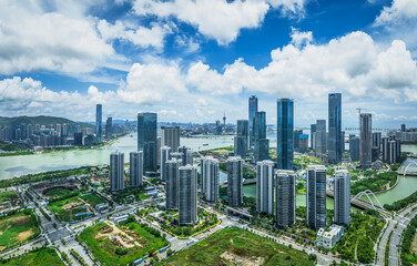 Obraz premium Expansive aerial cityscape view of a modern financial district with numerous skyscrapers and residential buildings alongside a river on a sunny day.