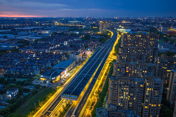 Fototapeta premium Modern elevated expressway illuminated with golden lights cutting through residential and industrial districts at twilight.