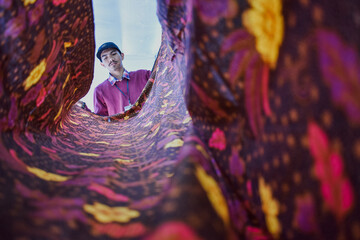 Low‑angle shot of a young craftsman inspecting a flowing sheet of vibrant Indonesian batik, its...