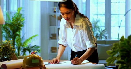 Indian female architect drafting drawing at desk in cozy modern office, Asian woman employee standing and manually sketching architectural plan using scale and pencil on paper with focused expression