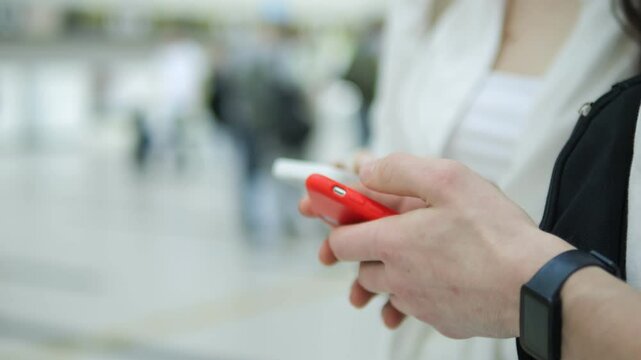 Man is holding a red cell phone in his hand. He is wearing a watch and a white shirt. There are other people in the background, some of them carrying handbags