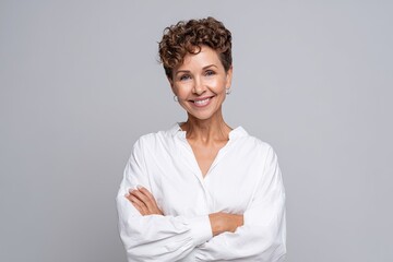 A woman with short curly hair in a white blouse standing with arms folded and smiling warmly The light gray