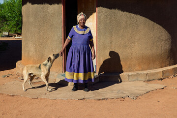 African village, single old woman with headscarf , in front of the house, dog playing