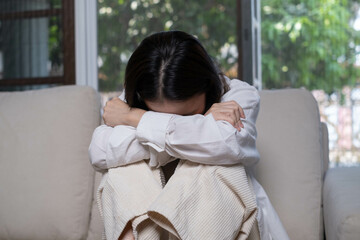 A woman sits on a couch, hugging her knees with a distressed expression, surrounded by natural...