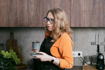 Blond woman in her kitchen prepares espresso using a capsule coffee machine and enjoys her coffee,...