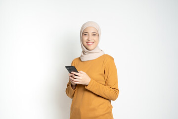Smiling woman wearing a hijab and mustard-colored sweater, holding a smartphone and posing...