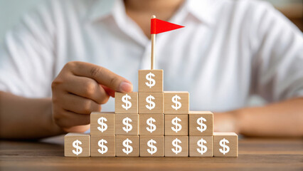 Concept of savings 2026. A person arranges wooden blocks with dollar signs into a pyramid, placing a red flag on top symbolizing financial success or goal achievement.