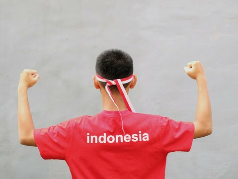 Indonesia young man in red t-shirt with INDONESIA written on it wearing red  white headband and clenched fist against grey background during Indonesian independence celebration, back view