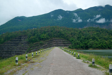A straight dam road flanked by posts and surrounded by a lake and dense forest, leading toward a misty green mountain under cloudy skies.