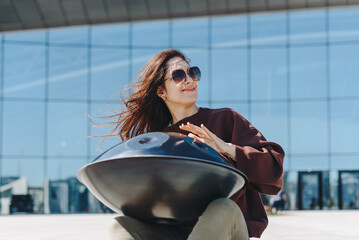 young caucasian woman playing black handpan with hands, ethnic metal drum instrument, sits on stone staircase, scyscraper and business center in background, beautiful relaxing music for meditation
