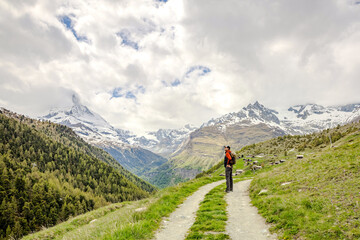 A lone hiker stands on a winding trail surrounded by mountains