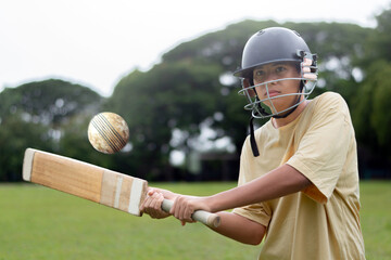 Young boy wearing a helmet plays cricket on a grassy field, focusing intensely as he hits the ball with a wooden bat. Outdoor sports and youth energy captured in motion.
