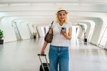 Young woman walking through a bright airport terminal, using her smartphone while pulling a suitcase, embodying the spirit of travel and adventure