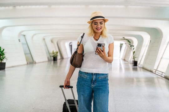 Young woman walking through a bright airport terminal, using her smartphone while pulling a suitcase, embodying the spirit of travel and adventure - Powered by Adobe