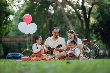 A family of four is sitting on a grassy field, playing music and eating fruit. A man is playing a...