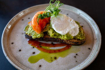 Close-up of avocado toast topped with a poached egg, smoked salmon, microgreens, and a drizzle of olive oil, served on artisan bread.