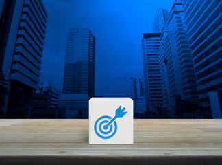 Target with dart flat icon on white block cube on wooden table over modern office city tower and...
