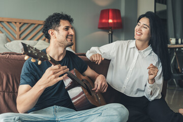 Happy young Caucasian couple bonding while sitting on floor with guitar, man playing while woman...