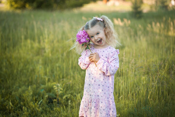cheerful 3-year-old girl with pigtails in a pink dress with a peony walks in a village meadow