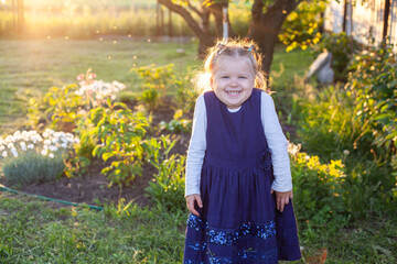 cheerful 3-year-old girl in a blue dress walks near the flowering flower bed in the backyard