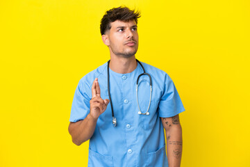 Young surgeon doctor man isolated on yellow background with fingers crossing and wishing the best