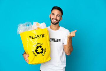 Young caucasian man holding a bag full of plastic bottles to recycle isolated on blue background...