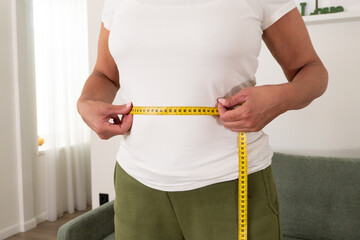 Mature woman measuring waist with tape at home, tracking size changes for healthy lifestyle, diet...