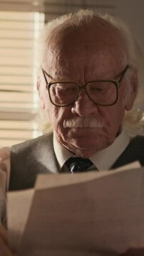 Vertical medium close up shot of focused senior Caucasian man in retro suit vest and eyeglasses studying paper documents sitting by window in office