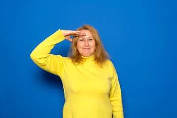 A smiling and proud Caucasian woman in her 40s saluting the camera in an act of honor and patriotism, showing respect. Isolated on a blue background