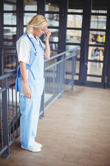 Nurse in scrubs leaning on railing holding forehead with stethoscope in mezzanine near glass doors