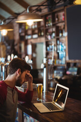 Laptop sitting on wooden bar counter beside pint of beer under industrial pendant lights in pub