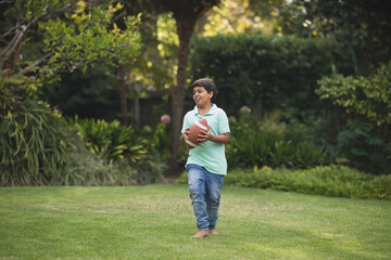 Hispanic child boy running barefoot on grass lawn in backyard garden holding brown football