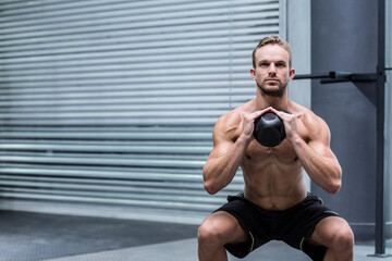 Shirtless man performing kettlebell squat at gym near metal roller shutter door on rubber flooring