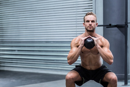 Shirtless man performing kettlebell squat at gym near metal roller shutter door on rubber flooring - Powered by Adobe