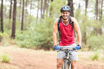 Man riding bicycle through pine forest wearing helmet, red tank top, blue gloves, carrying backpack