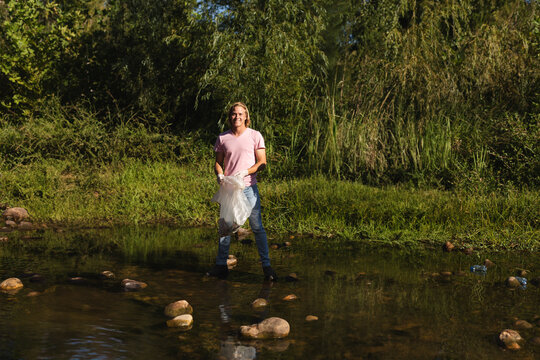 Male volunteer wearing knee-high boots collecting plastic bottles in shallow creek among stones