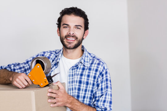 Man sealing cardboard box with orange tape dispenser while standing in empty room